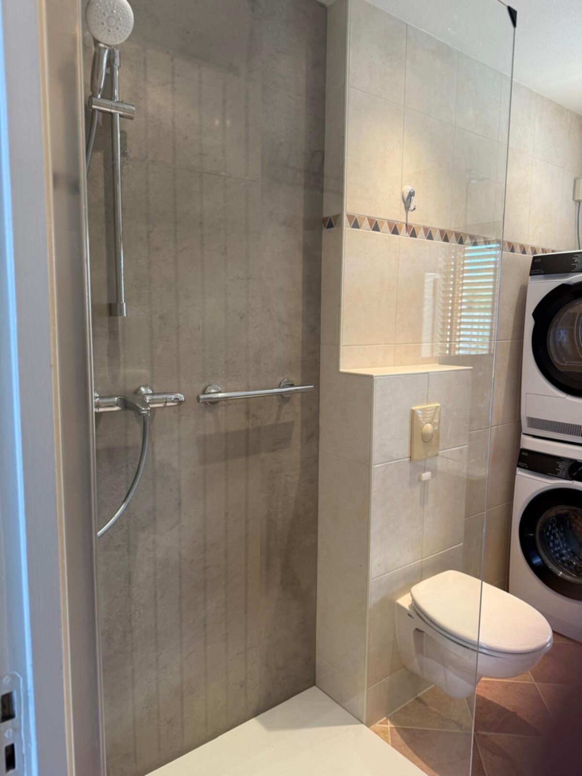 Douche en toilet in kleine badkamer met grijze en beige tegels en witte wasmachines
