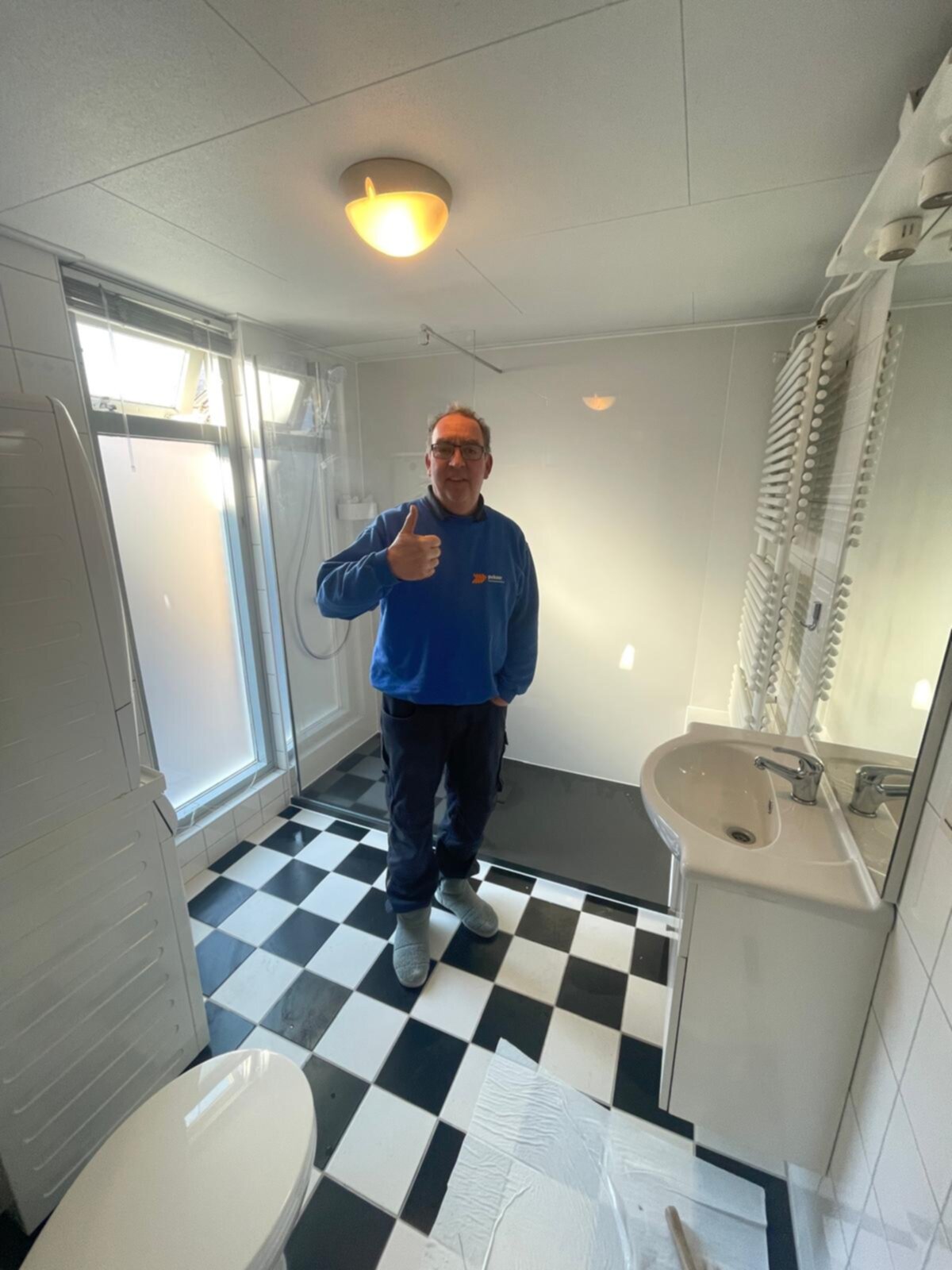 Een man in een blauwe trui geeft duim omhoog in een kleine badkamer met zwart-wit betegelde vloer