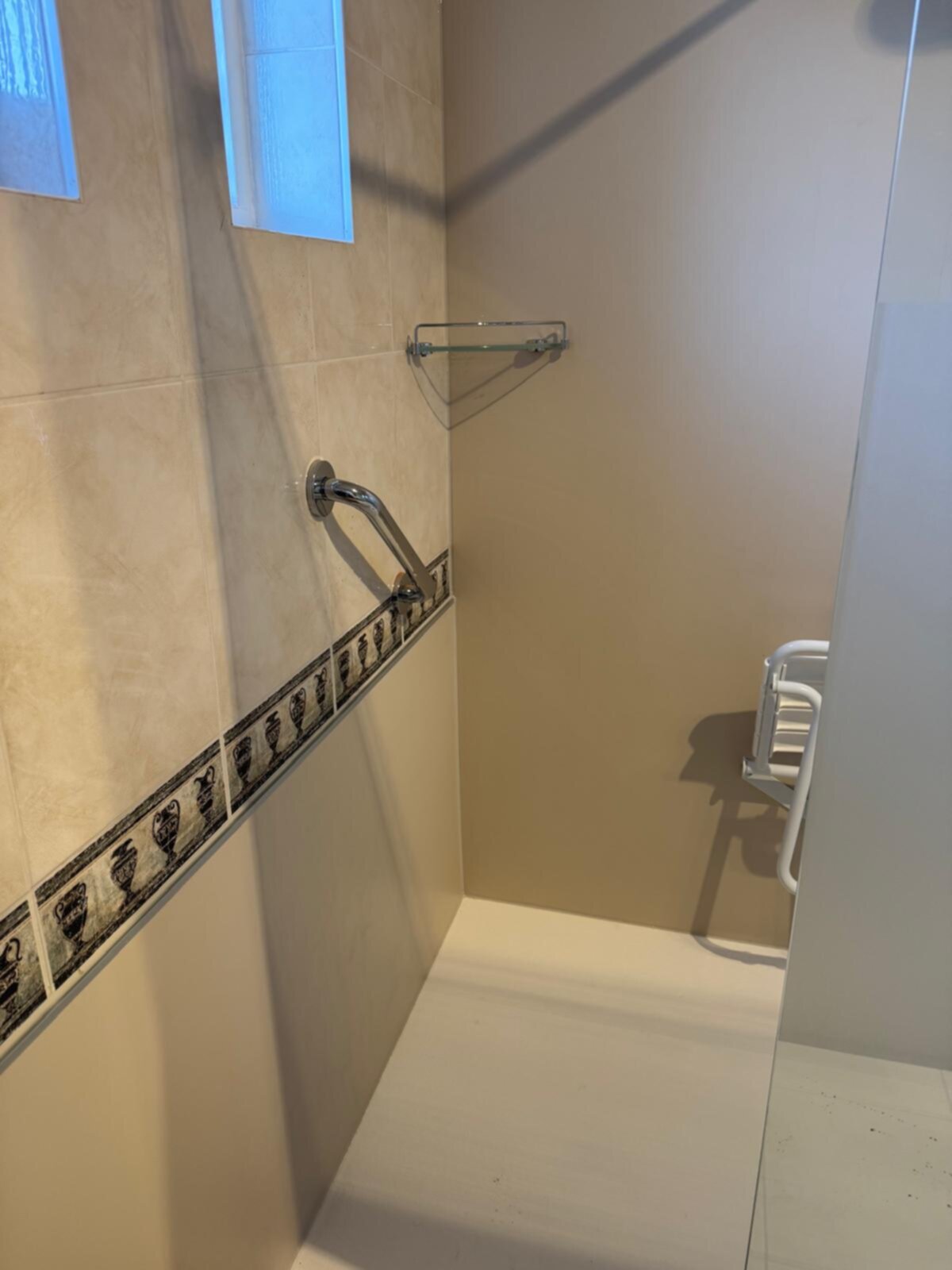 Douchehoek met beige tegels en decoratieve rand, chroom handgreep en glas shelf.