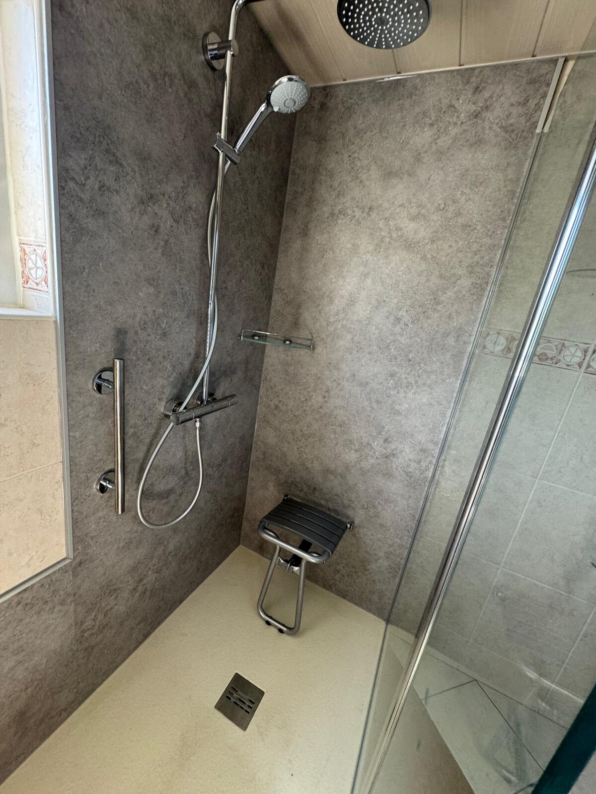 Douche met grijze wanden en beige vloer, toegankelijk met stoel en glazen deur