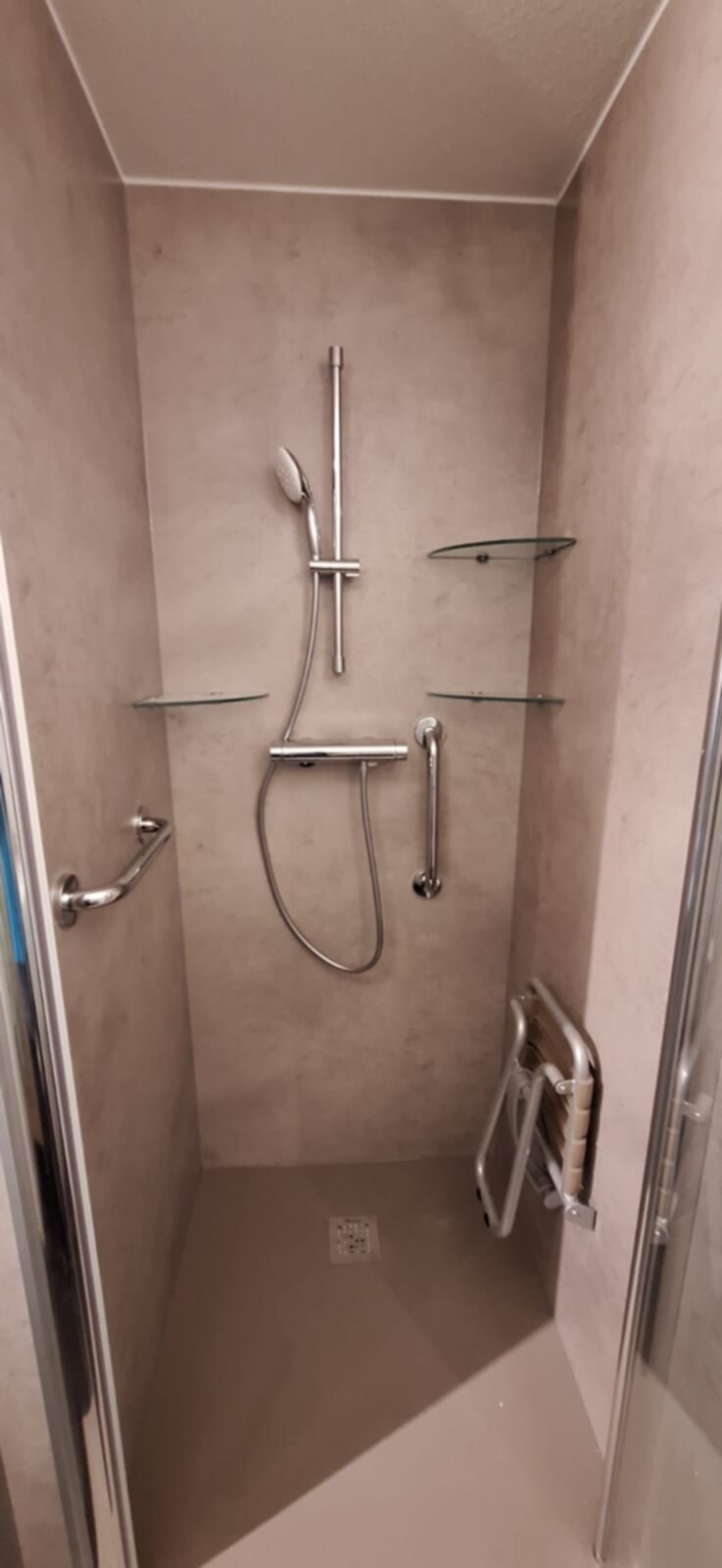 Douchecabine met beige betegeling, glazen planken, metalen douchekop en grepen.