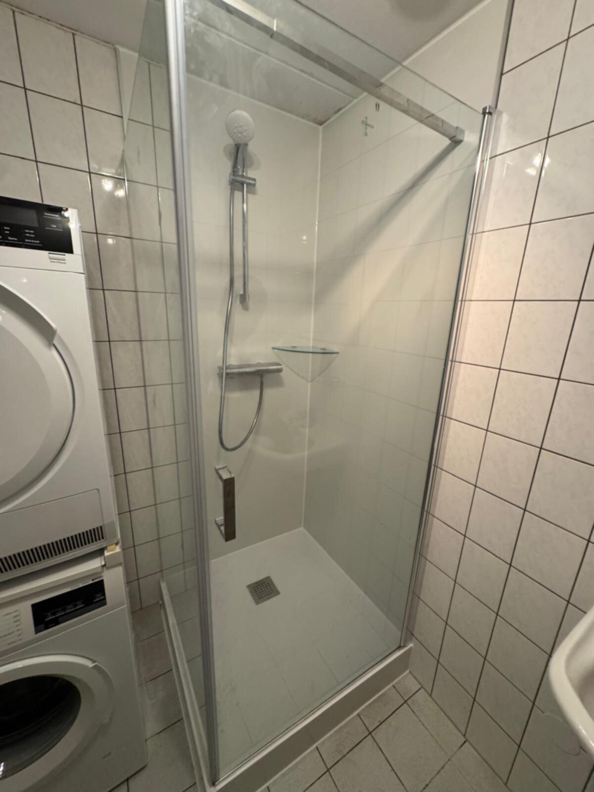Douchecabine met witte tegelwanden en glazen deur, metalen douchekop en wasmachine