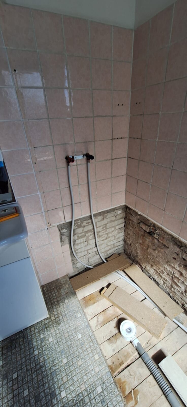 Slechte staat roze betegelde badkamer met opengelegde leidingen en houten vloerdelen