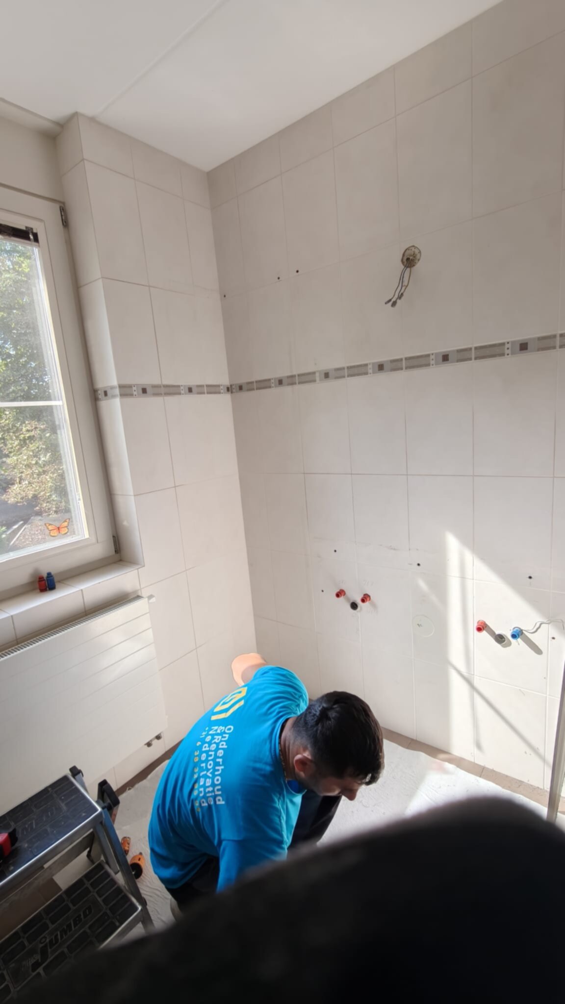 Wit betegelde badkamer met werker in blauwe trui tijdens renovatie