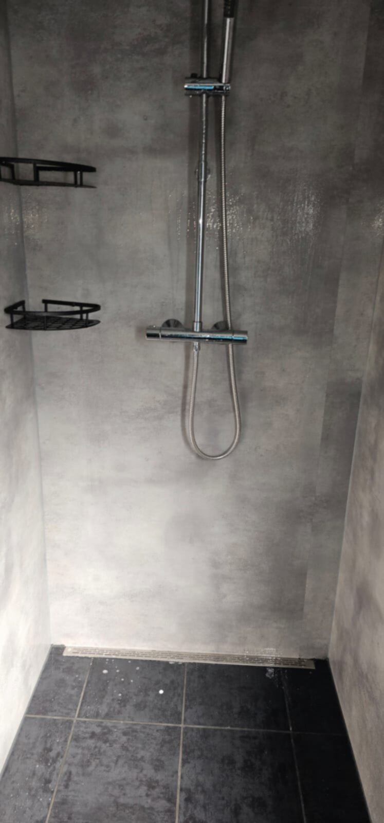 Douche met grijs betegelde wanden en zwarte vloer, metalen douchekop en twee zwarte rekken.