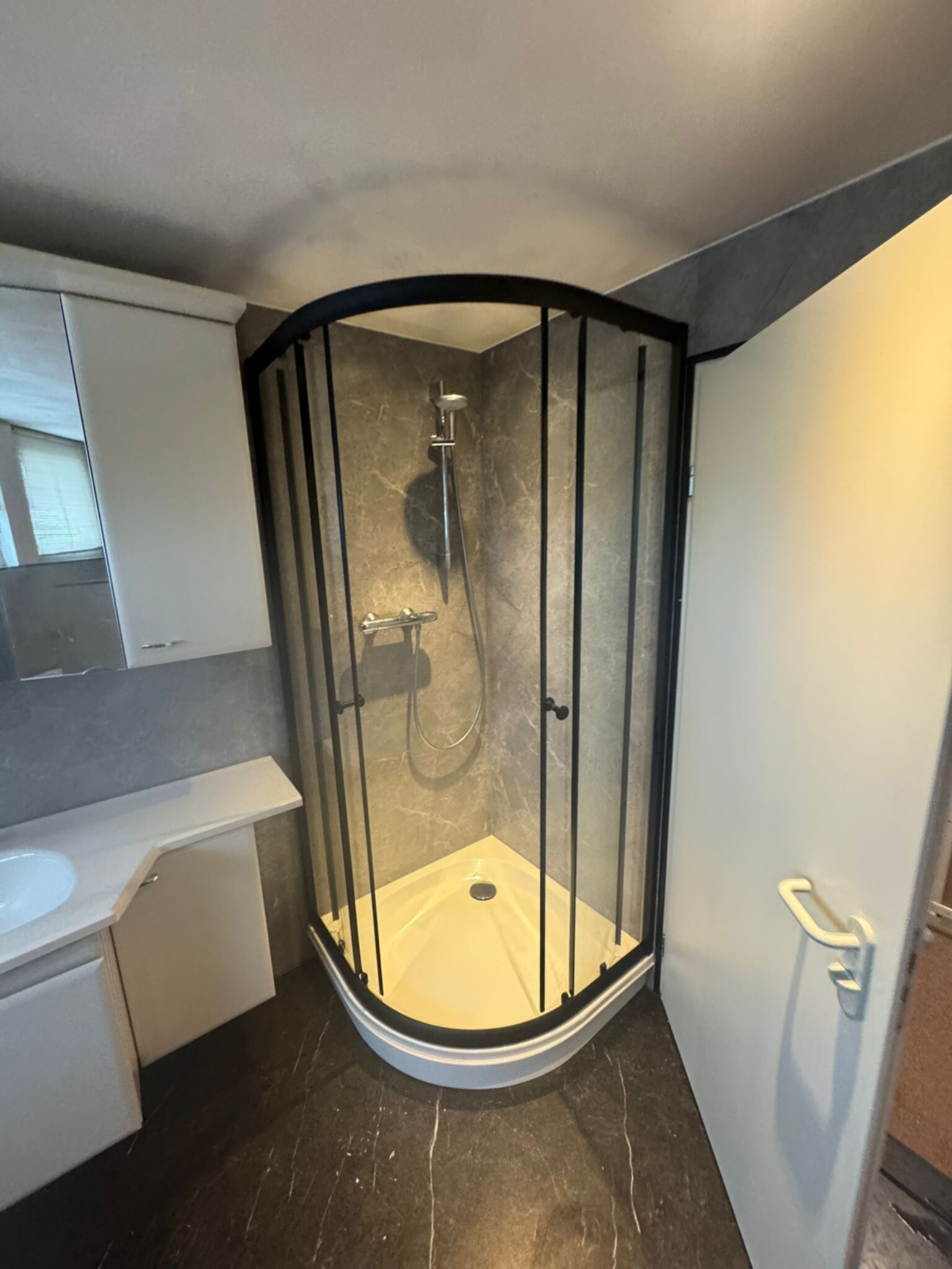 Badkamer met grijze tegel, witte douche en donkere vloer